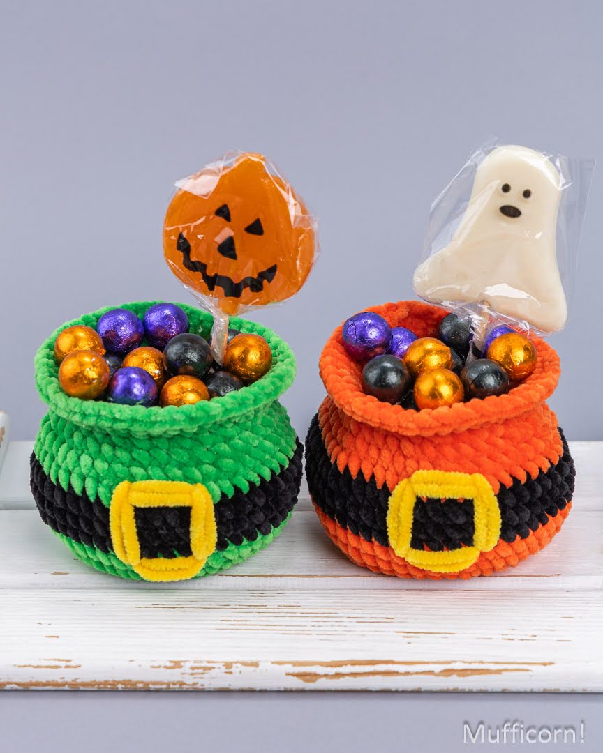 Two crocheted Halloween pot holders filled with colorful foil candies, with a pumpkin-shaped lollipop in one and a ghost-shaped candy in the other, displayed on a white wooden surface against a gray background.