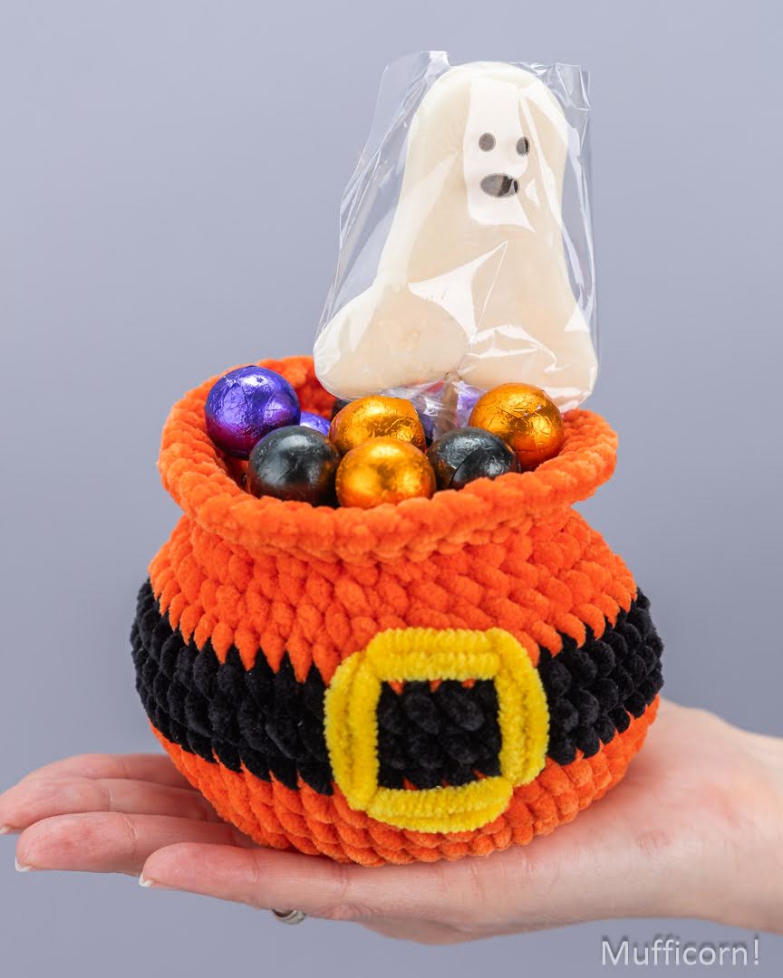 A crocheted orange and black pot with a yellow buckle detail, filled with colorful foil candies and topped with a ghost-shaped lollipop, held in a person's hand against a plain gray background.