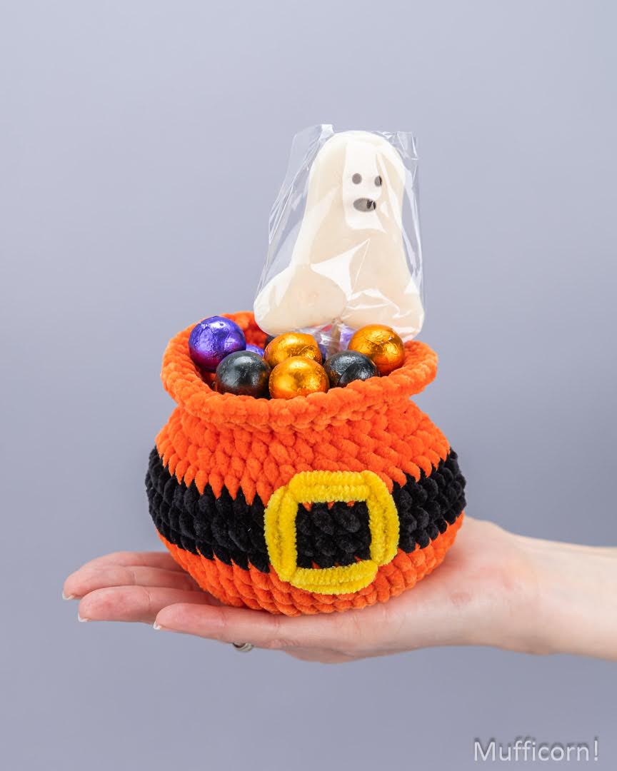 A crocheted orange and black pot-shaped candy holder with a yellow buckle detail, held in a person's hand. The pot is filled with colorful chocolate eggs and a ghost-shaped candy under clear plastic, against a plain gray background.