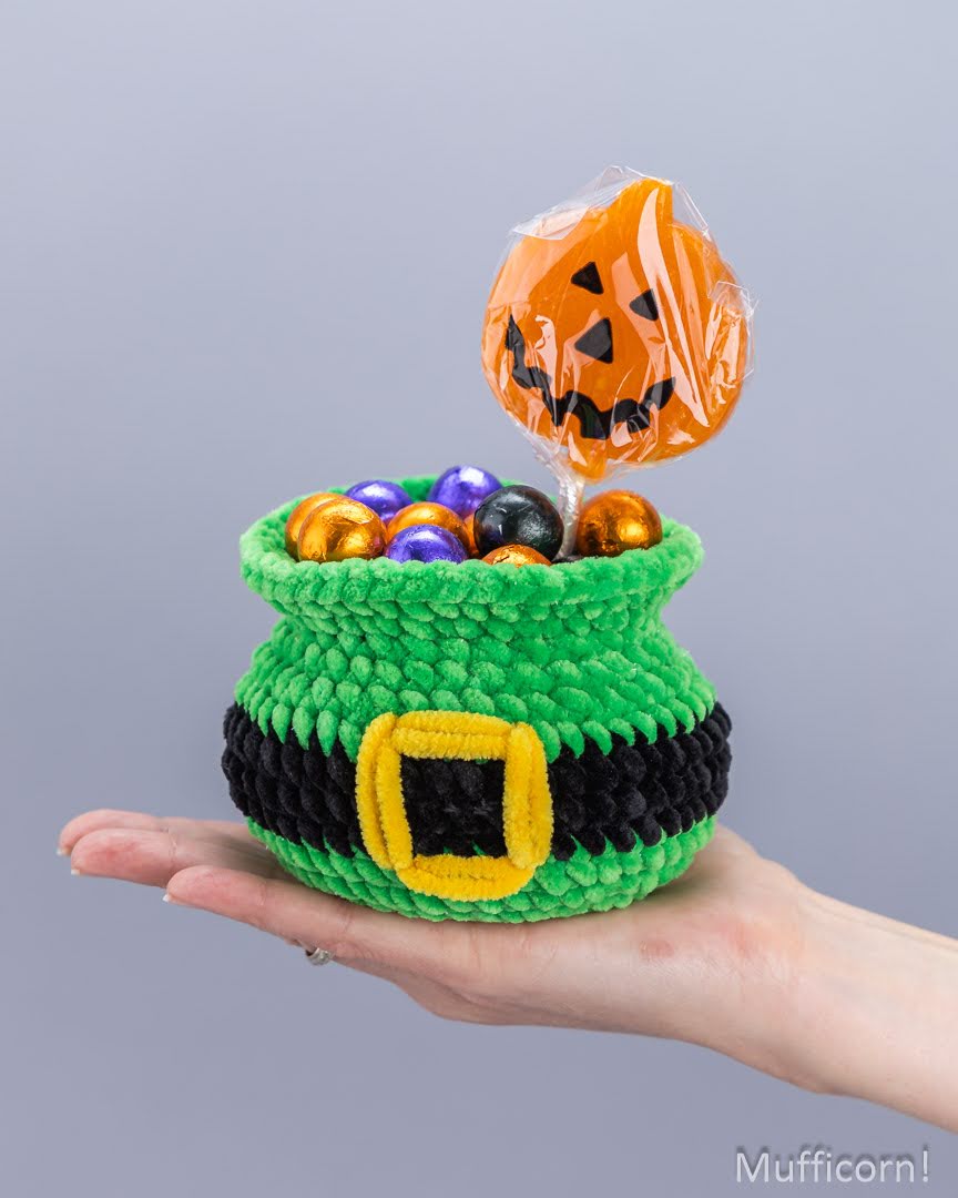 A hand holds a small crocheted green pot with a black and yellow belt detail, filled with colorful wrapped candies, with an orange jack-o'-lantern lollipop sticking out against a plain gray background.
