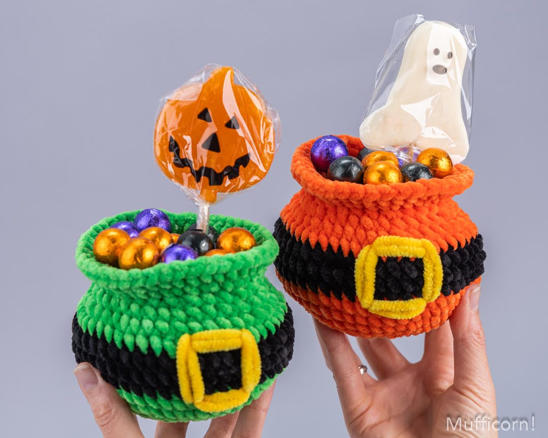 Two handmade crocheted Halloween pot holders being held in hands against a solid gray background. The green pot has a jack-o'-lantern candy holder, and the orange pot has a ghost candy holder.