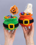 Two hands holding crocheted Halloween-themed candy pots filled with colorful foil candies, with a jack-o'-lantern lollipop in one pot and a white ghost lollipop in the other, against a plain gray background.