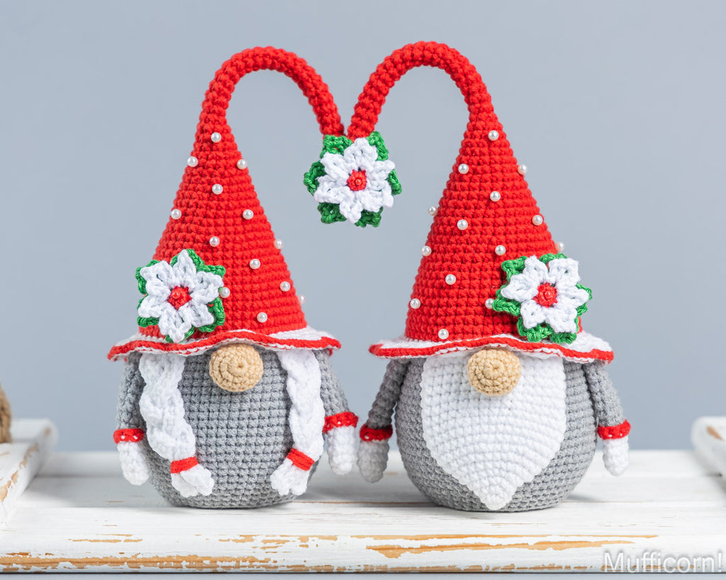 Two crocheted Christmas gnomes with red pointed hats, white beards, gray bodies, and white flower ornaments, standing on a white wooden surface against a light gray background.