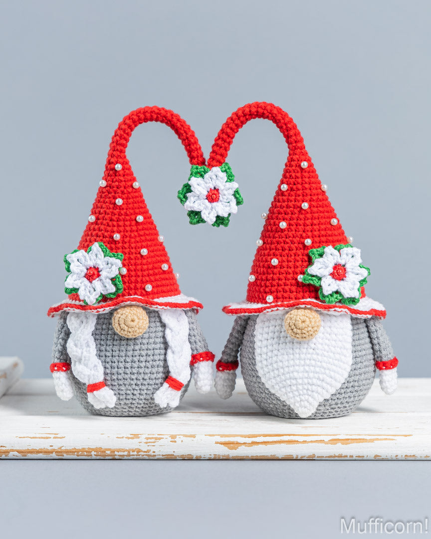 Two crocheted Christmas gnomes standing side by side on a white rustic surface against a light gray background. Each gnome has a red pointed hat with white polka dots, a white flower with green leaves, a gray body, and a white beard.