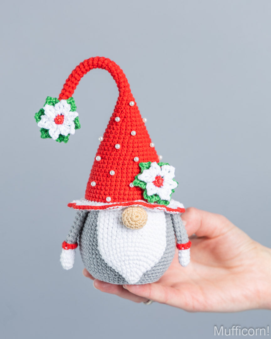 A crocheted Christmas gnome doll with a red pointed hat, white beard, gray body, and white flowers, held in a person's hand against a plain gray background.
