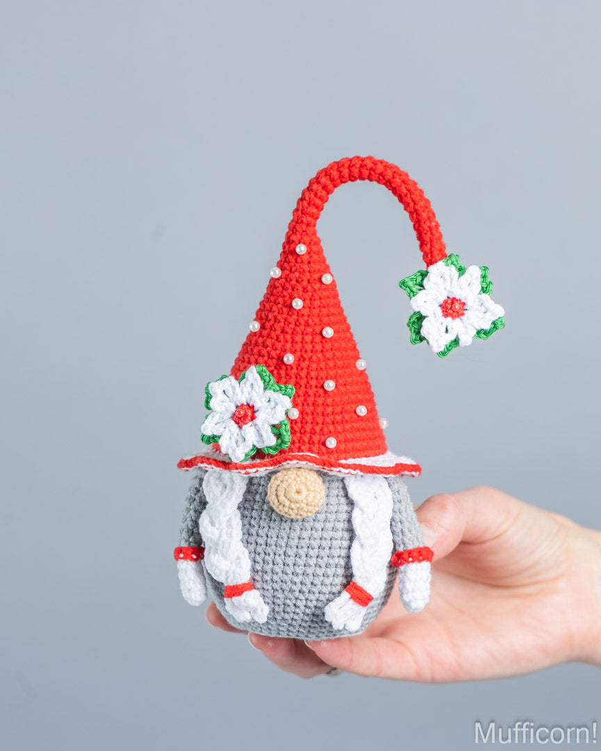 A crocheted gnome ornament held in a person's hand against a plain light gray background. The gnome has a red pointed hat with white polka dots and white flower accents, a gray body, white braided hair, and a small beige snout.