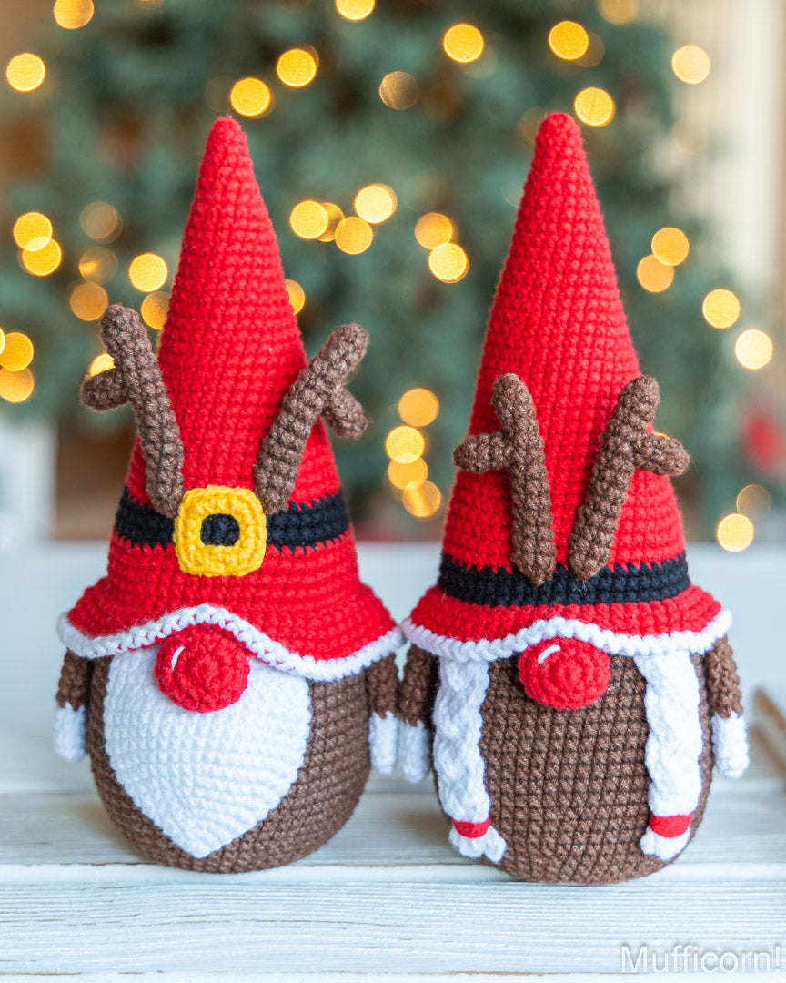 Two crocheted holiday gnome ornaments styled as reindeer, featuring red pointed hats with brown antlers, black and yellow belts, white beards, and brown bodies, displayed on a white wooden surface with a blurred Christmas tree and warm lights in the background.