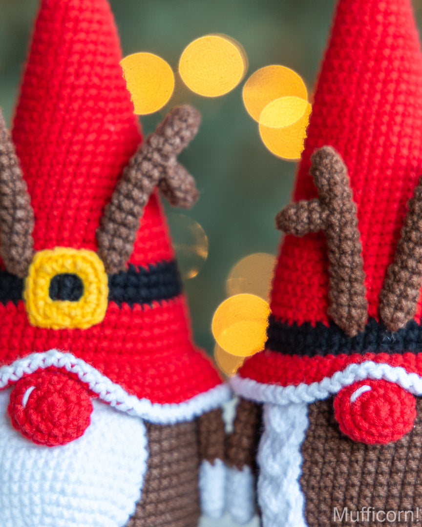 Close-up of two crocheted Christmas gnome ornaments with red pointed hats, brown antlers, black belts, yellow buckle details, white and brown bodies, and a festive blurred holiday background.