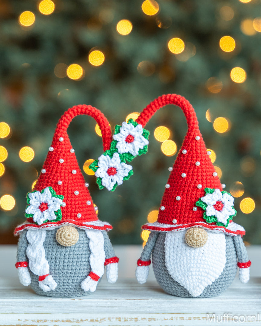 Two crocheted Christmas gnomes with red pointed hats, white braids, gray bodies, and white flower decorations, posed together on a light surface with a blurred holiday tree and warm lights in the background.