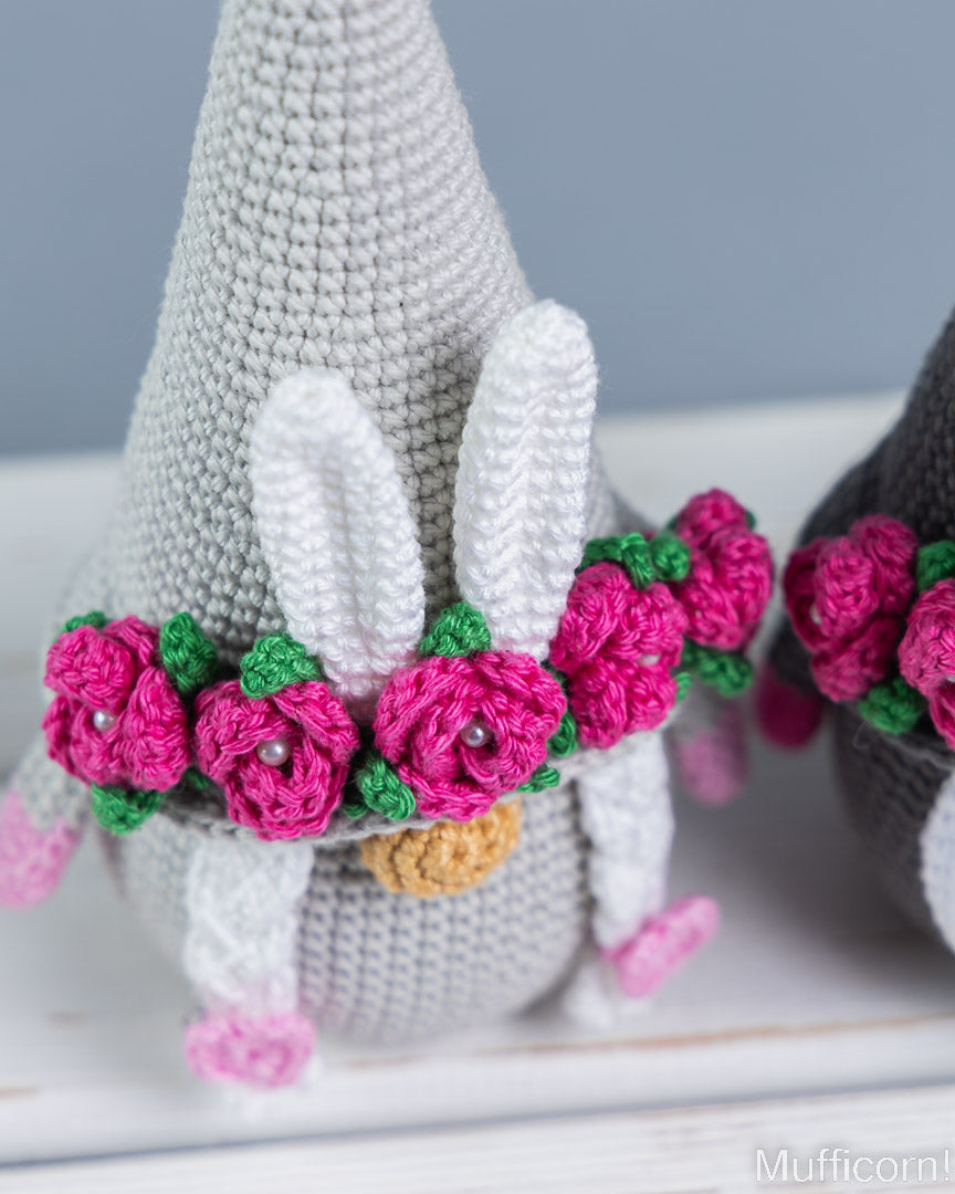 A crocheted bunny-shaped ornament or doll with a gray body, white ears, a pink flower crown with green leaves, and a small yellow beak, shown on a white surface against a soft blue-gray background.