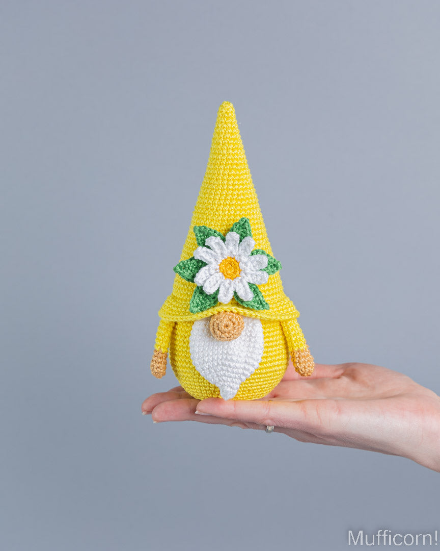 A crocheted gnome doll with a tall yellow hat, white beard, and daisy decoration is being held in a person's hand against a plain gray background.