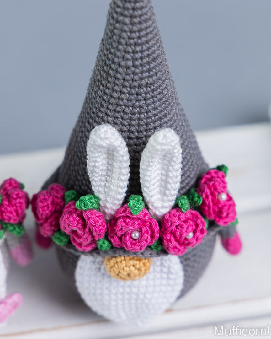 A crocheted bunny gnome decoration with a gray pointed hat, white ears, a white face, pink flowers with green leaves, and a small gold nose, shown close up on a white surface.