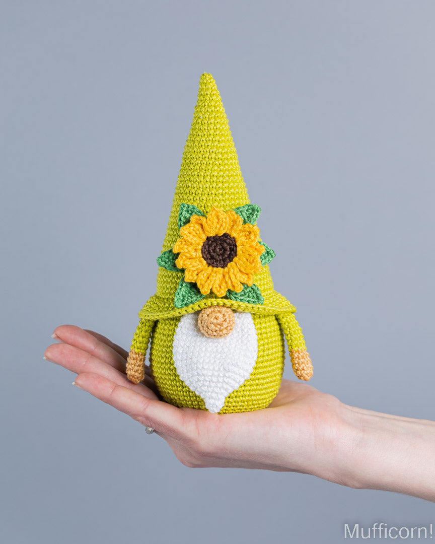 A crocheted gnome doll with a tall yellow hat, sunflower face, green leaves, white beard, and tan nose is being held in a person's hand against a plain gray background.