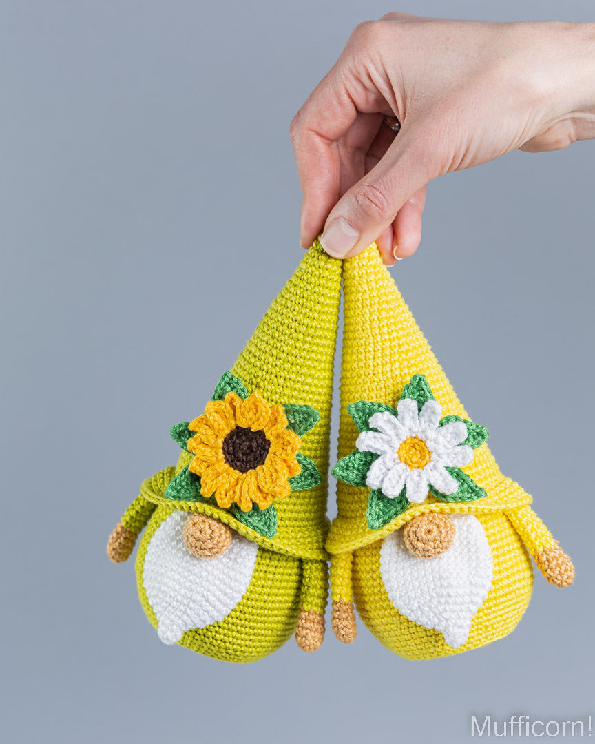 A hand holds two crocheted gnome figures with pointed yellow hats decorated with a sunflower and a white daisy, against a plain gray background.