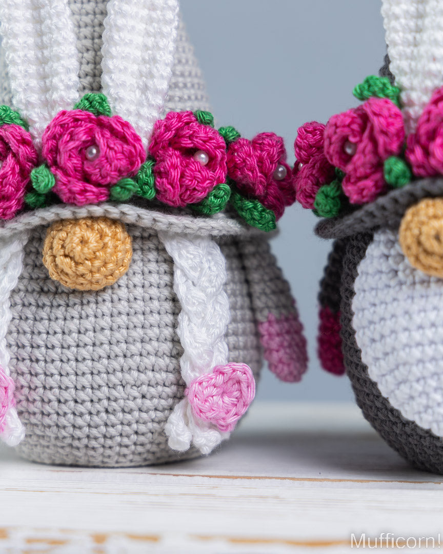 Close-up of crocheted bunny hats in gray and white with bright pink flowers and green leaves, displayed on a light surface against a soft gray background.