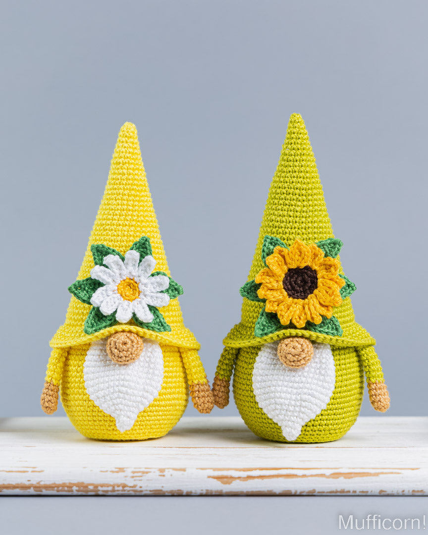 Two crocheted gnome figurines standing side by side on a white wooden surface against a light gray background. The left gnome has a white daisy on its hat, and the right gnome has a yellow sunflower on its hat.