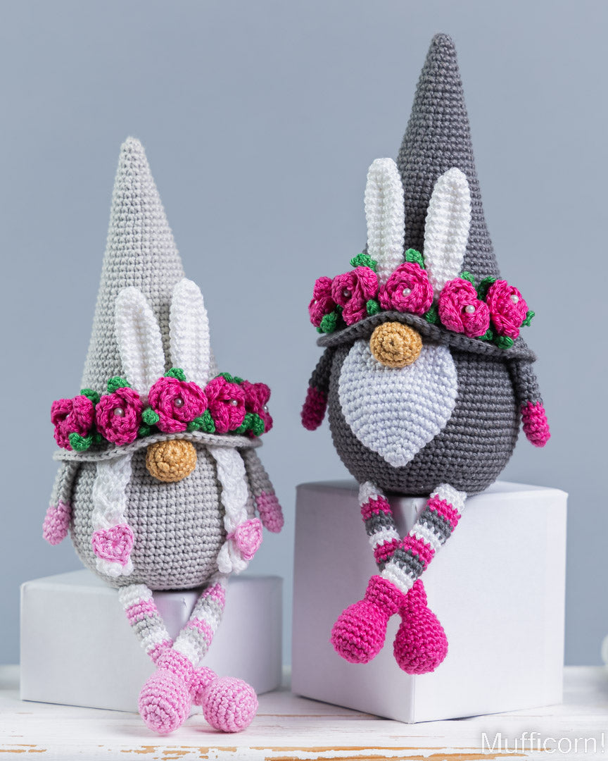 Two crocheted bunny gnome decorations sitting on white blocks against a light gray background. One is light gray and the other is dark gray, both wearing pink flower crowns, white bunny ears, striped socks, and pink shoes.