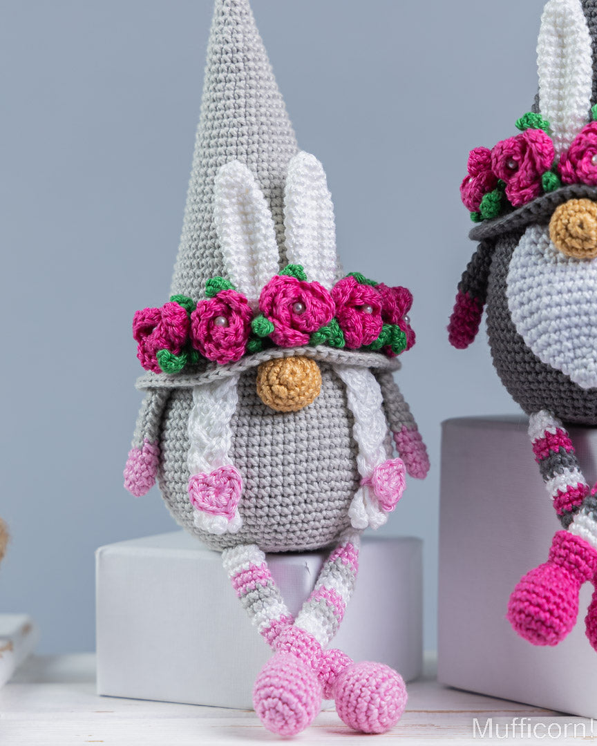 A crocheted bunny gnome doll with a tall gray hat, pink flower crown, and pink-and-white striped legs, posed on a light gray pedestal. A second similar doll is partially visible on the right.