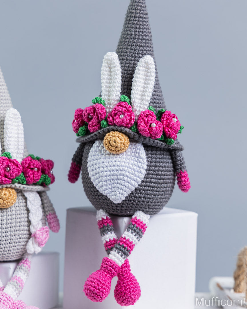 Crocheted bunny gnomes with gray pointed hats, white beaks and inner ears, pink flower crowns, striped pink/gray/white legs, and bright pink boots, displayed on white pedestals against a soft light gray background.