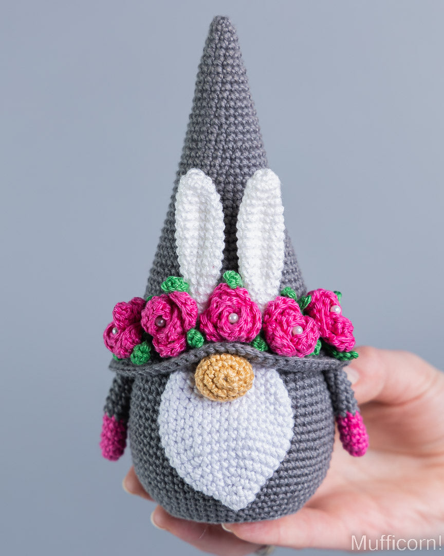 A crocheted gnome doll with a tall gray hat, white bunny ears, a pink flower crown, and a white chest patch, being held in a person's hand against a plain gray background.