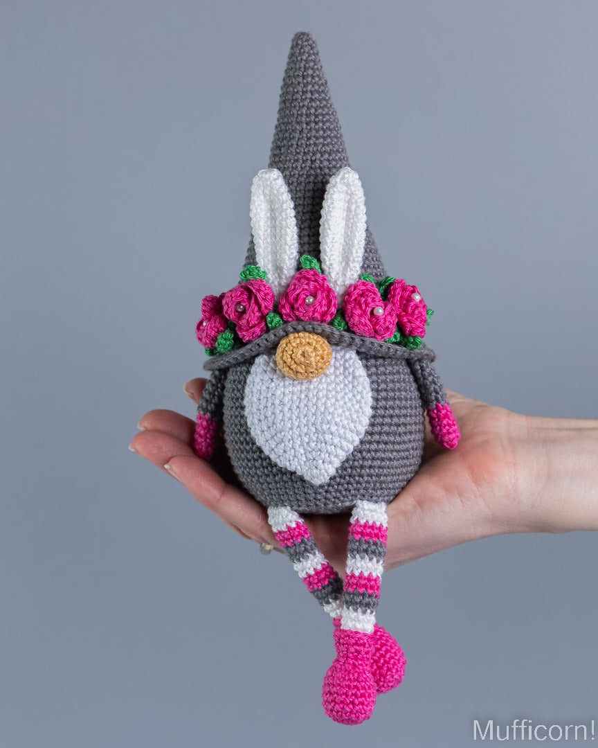 A crocheted bunny gnome doll with a gray pointed hat, white face and chest, pink flowers, and pink-and-white striped legs, being held in a person's hand against a plain gray background.