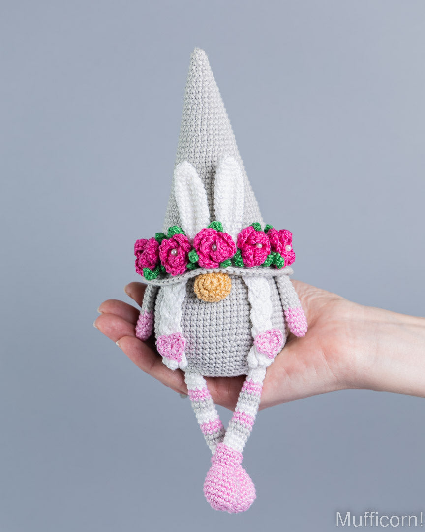 A crocheted bunny gnome doll with a tall gray hat, white inner ears, a pink flower crown, and striped legs, being held in a person's hand against a plain gray background.