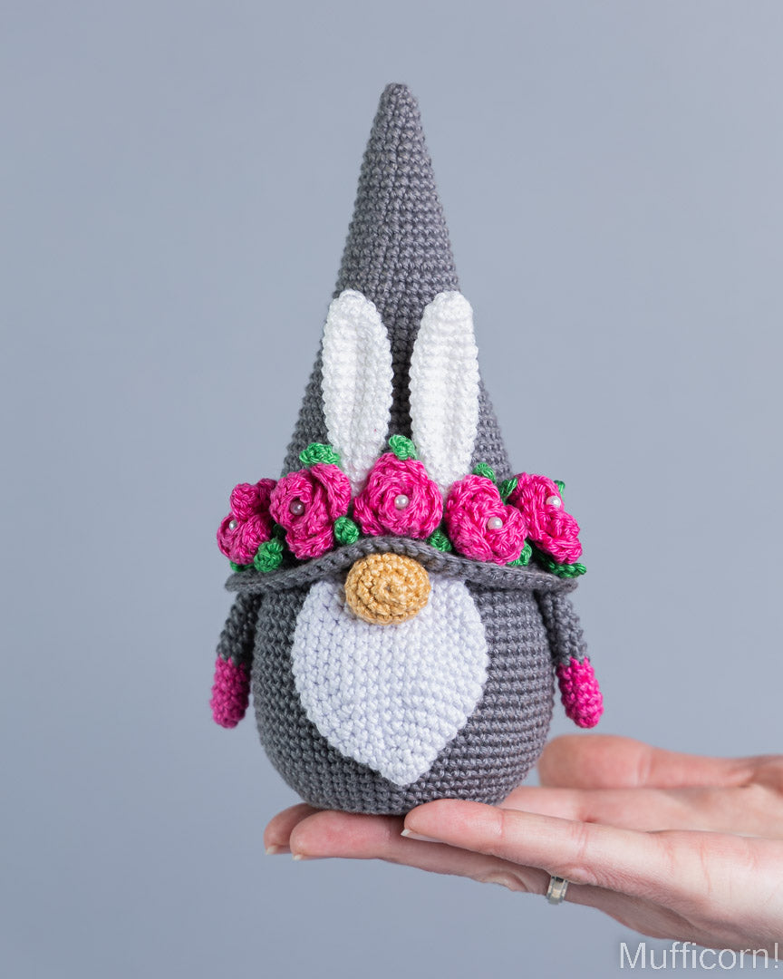 A crocheted bunny gnome doll with a gray body, white face and ears, pink flowers, and a gold snout, being held in a person's hand against a plain gray background.