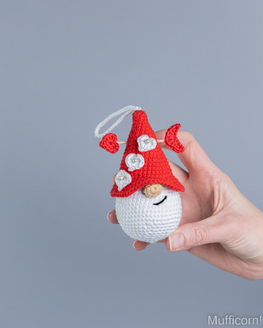 A hand holding a small crocheted gnome ornament against a solid gray background. The gnome has a red pointed hat with white flower details, a white beard, and a black stitched smile.