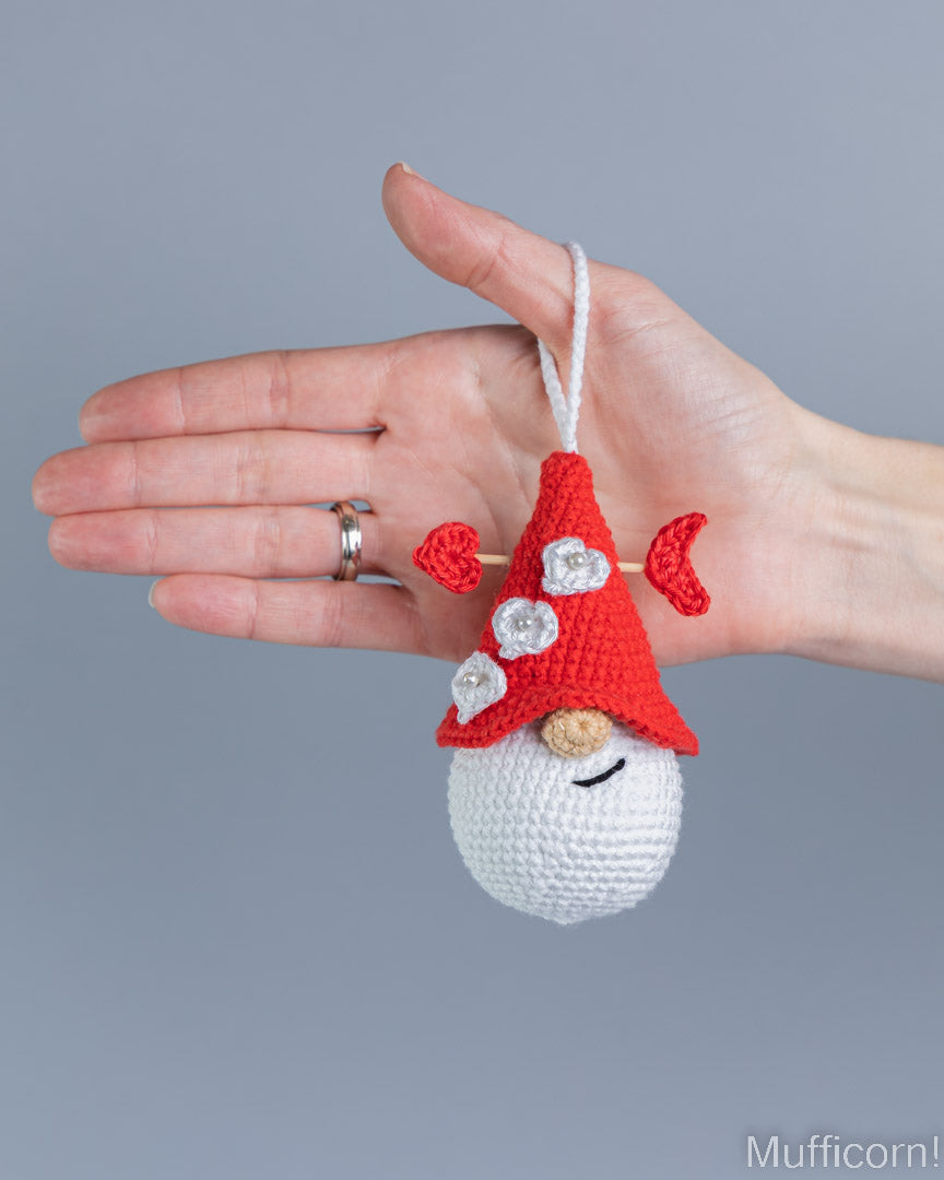 A small crocheted gnome ornament with a red pointed hat, white beard, and silver rhinestone accents, held in a person's hand against a plain gray background.