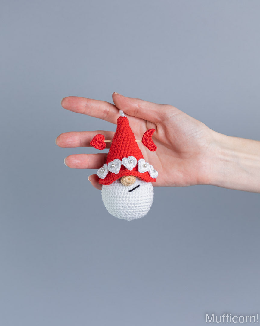 A hand holding a small crocheted gnome ornament with a red hat, white beard, and red heart details against a plain gray background.
