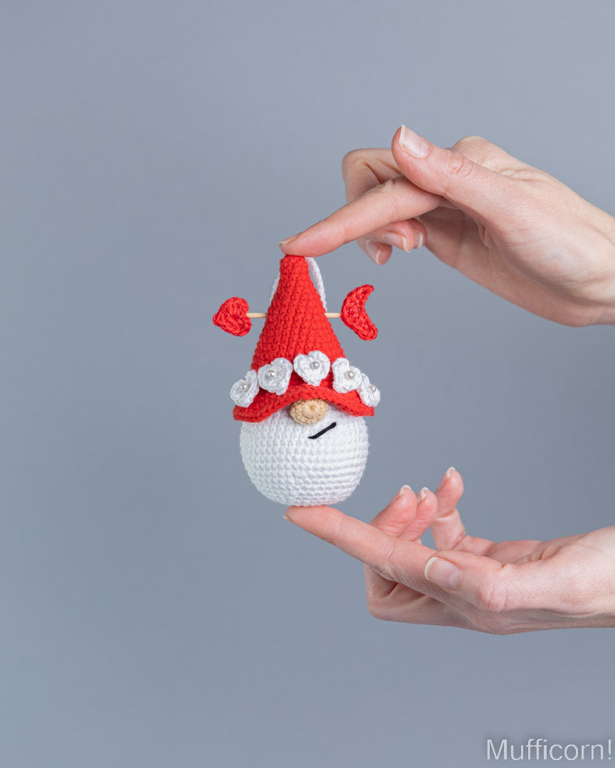 A small crocheted gnome doll with a red hat, white beard, and embroidered flowers is held between two human hands against a plain gray studio background.