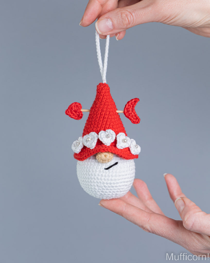 A small crocheted gnome ornament with a red pointed hat, white beard, and white flower trim is being held up by a person's hand against a plain gray background.