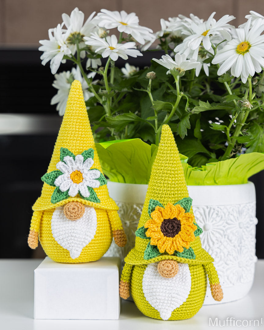 Two crocheted garden gnome decorations in bright yellow and chartreuse green, styled in front of a white pot of daisies on a white surface.