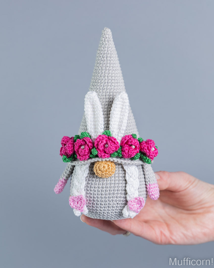 A crocheted bunny gnome doll with a gray body, white pointed hat, pink flowers, green leaves, and a gold nose, held in a person's hand against a plain gray background.