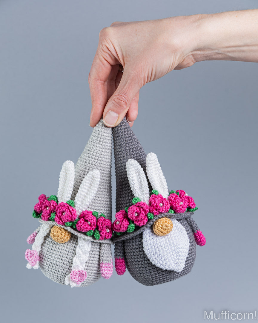 A hand holds two crocheted bunny-shaped muffs against a plain gray background. One bunny is light gray with white ears and a pink flower crown, and the other is dark gray with a white face and matching pink flowers.