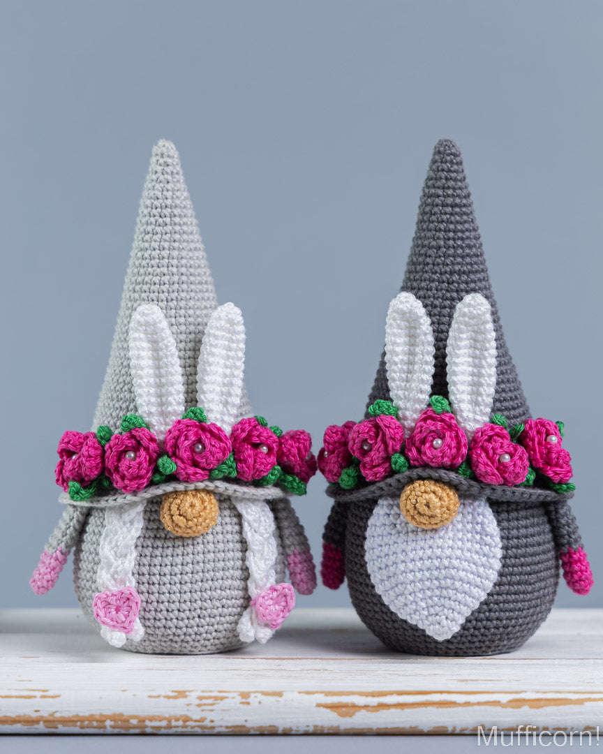 Two crocheted gnome figures with pointed hats and bunny ears, one light gray and one dark gray, both decorated with pink flower crowns and standing on a white rustic surface against a blue-gray background.