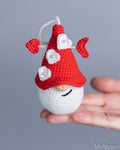 A small crocheted gnome ornament held in a person's hand against a soft gray background. The ornament has a red hat with white flower details, a white body, and a black stitched smile.