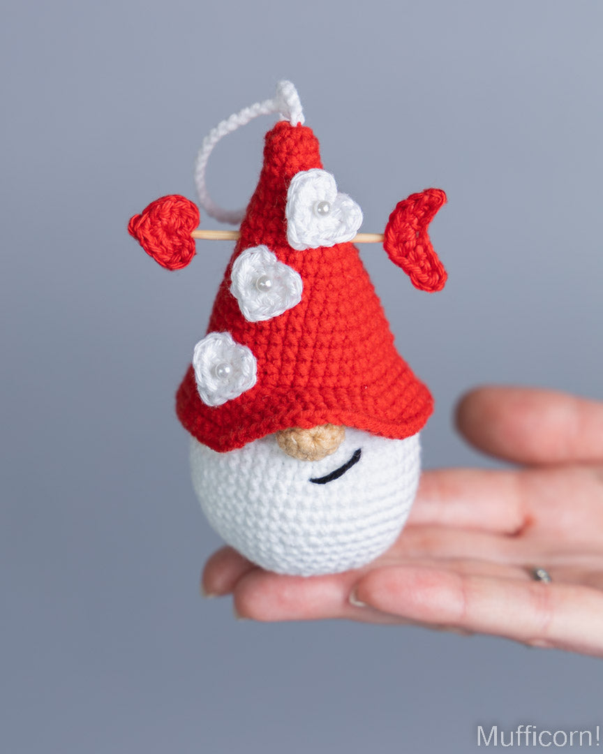 A small crocheted gnome ornament held in a person's hand against a soft gray background. The ornament has a red hat with white flower details, a white body, and a black stitched smile.