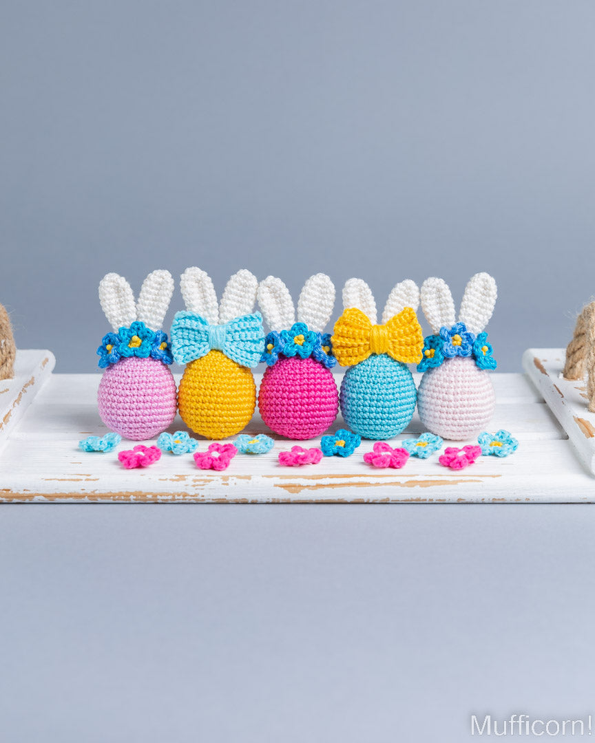Four crocheted Easter egg bunnies arranged on a white rustic board against a soft gray background. The bunnies have white yarn ears, pastel pink, yellow, magenta, and light blue bodies, blue flower crowns, and yellow or blue bows.