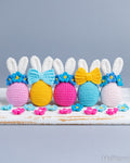 Five crocheted Easter bunny eggs lined up on a rustic white wooden surface against a solid gray background. The eggs are pink, yellow, and blue, each decorated with a matching bow and white yarn ears, accented with small blue and pink flowers.