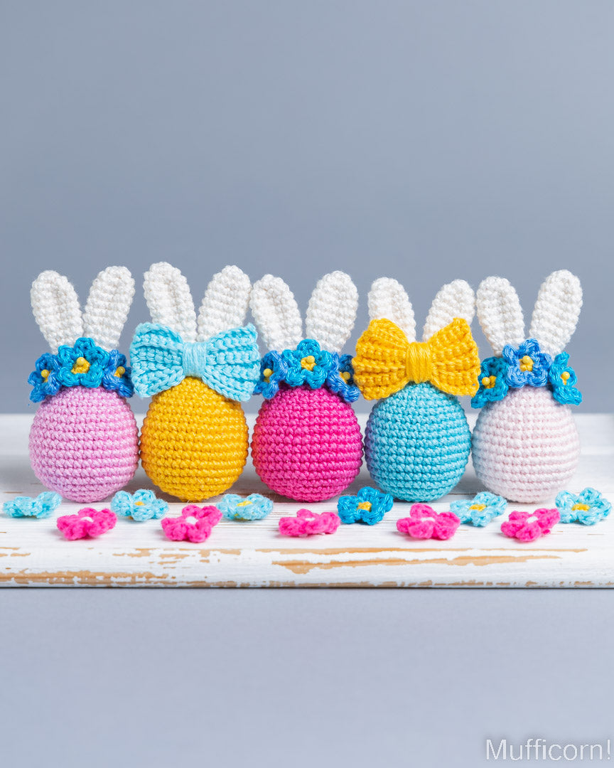 Five crocheted Easter bunny eggs lined up on a rustic white wooden surface against a solid gray background. The eggs are pink, yellow, and blue, each decorated with a matching bow and white yarn ears, accented with small blue and pink flowers.