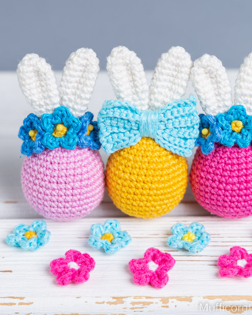 Three crocheted Easter egg characters with bunny ears, arranged on a white wooden surface. The center egg is yellow with a light blue bow, flanked by pink and magenta eggs with blue flowers.
