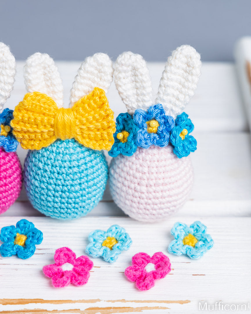 Three crocheted bunny eggs in pastel pink, light blue, and magenta, decorated with white yarn ears, a yellow bow, and blue flower crowns, arranged on a white wooden surface with loose flower embellishments in front.