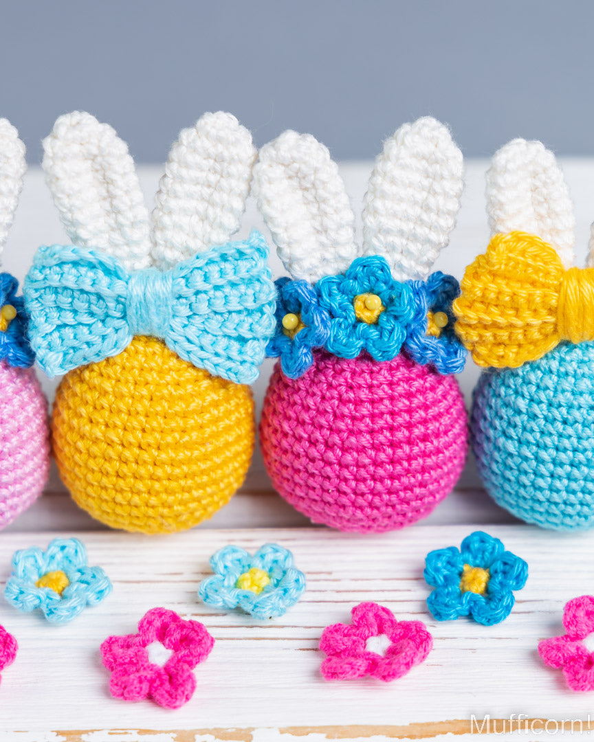 Three crocheted Easter eggs shaped like bunnies, decorated with white bunny ears, blue bows, blue flower crowns, and yellow and blue yarn bodies, arranged on a white wooden surface with small matching flowers in front.