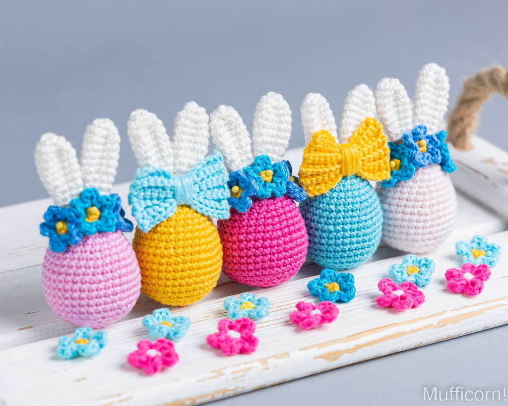 Five crocheted Easter eggs shaped like bunnies, each with white yarn ears, a colorful bow, blue flower accents, and pastel egg bodies in pink, yellow, magenta, light blue, and lavender, arranged on a white wooden surface with small matching flowers in front.