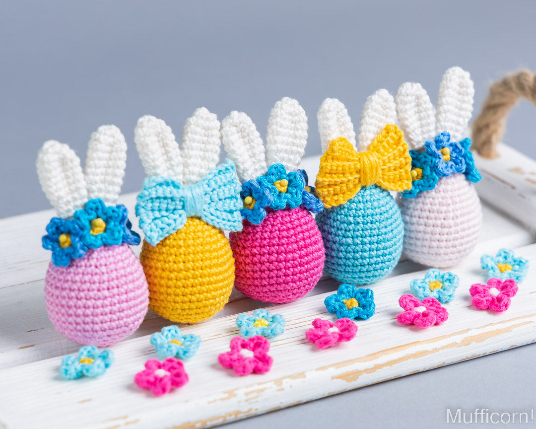 Five crocheted Easter eggs shaped like bunnies, each with white yarn ears, a colorful bow, blue flower accents, and pastel egg bodies in pink, yellow, magenta, light blue, and lavender, arranged on a white wooden surface with small matching flowers in front.