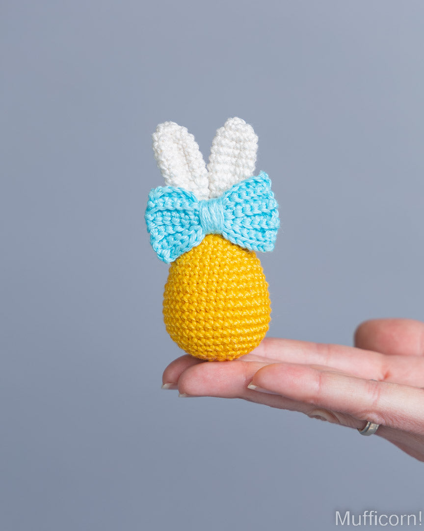 A small crocheted yellow egg ornament with white bunny ears and a light blue bow, held in a person's hand against a plain gray background.