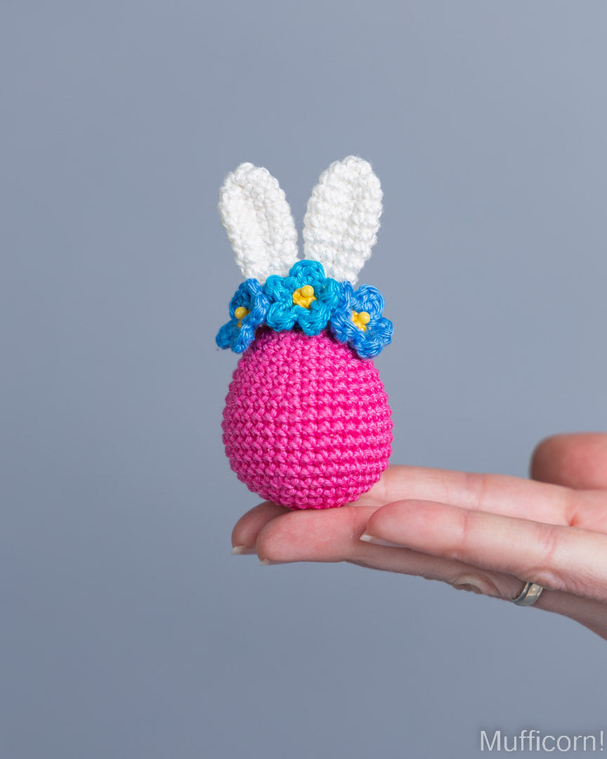 A small crocheted bunny egg decoration in bright pink with white bunny ears, a blue flower crown, and yellow flower centers, being held on a person's hand against a plain gray background.