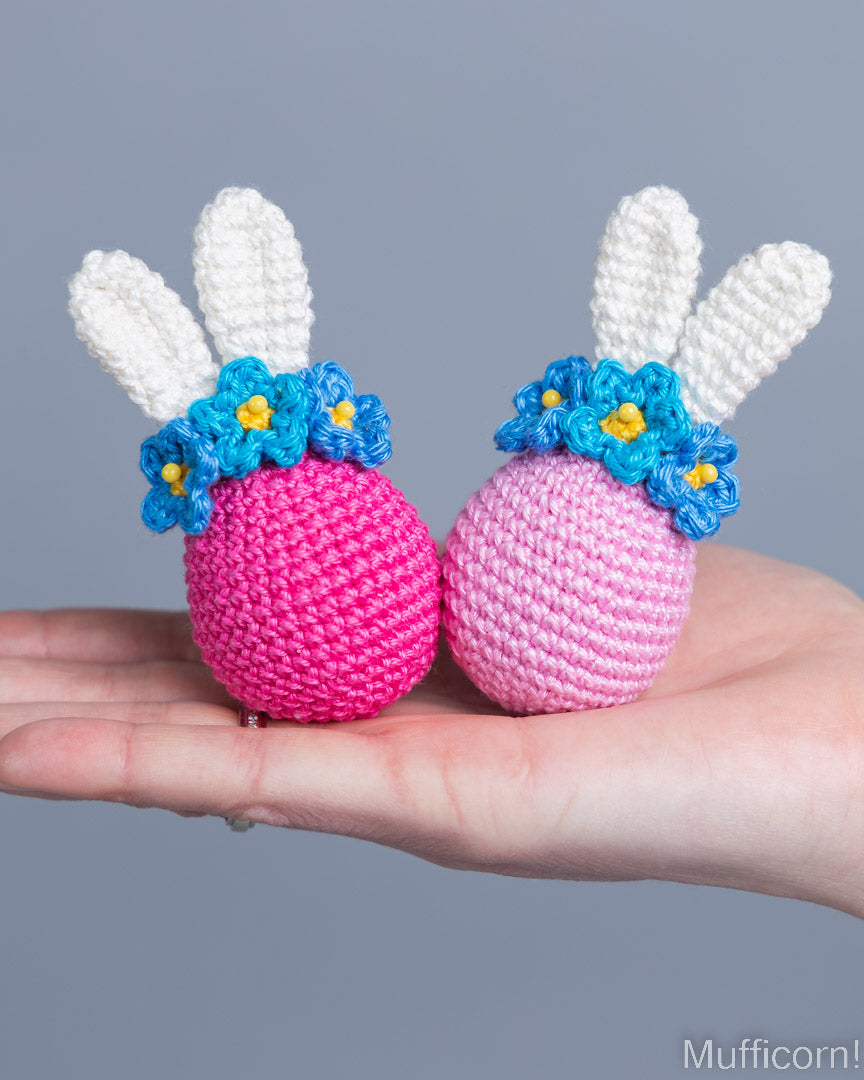 Two crocheted bunny Easter eggs held in a person's hand against a solid gray background. The eggs are pink and light pink, decorated with white yarn bunny ears and blue flower crowns with yellow centers.