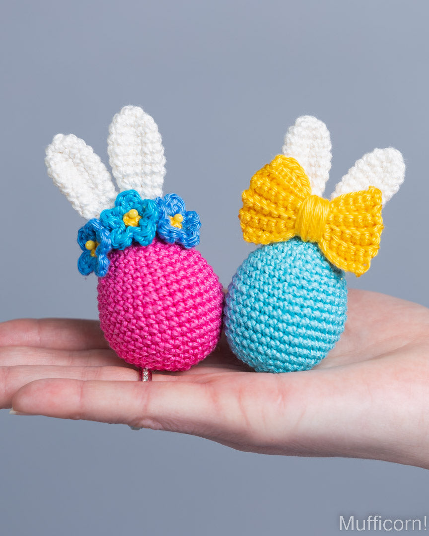 Two crocheted bunny toys, one pink with blue flowers and one blue with a yellow bow, held in a hand against a gray background.